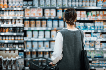 Donna di spalle fa la spesa al supermercato scegliendo prodotti dagli scaffali pieni di latte in polvere e alimenti.