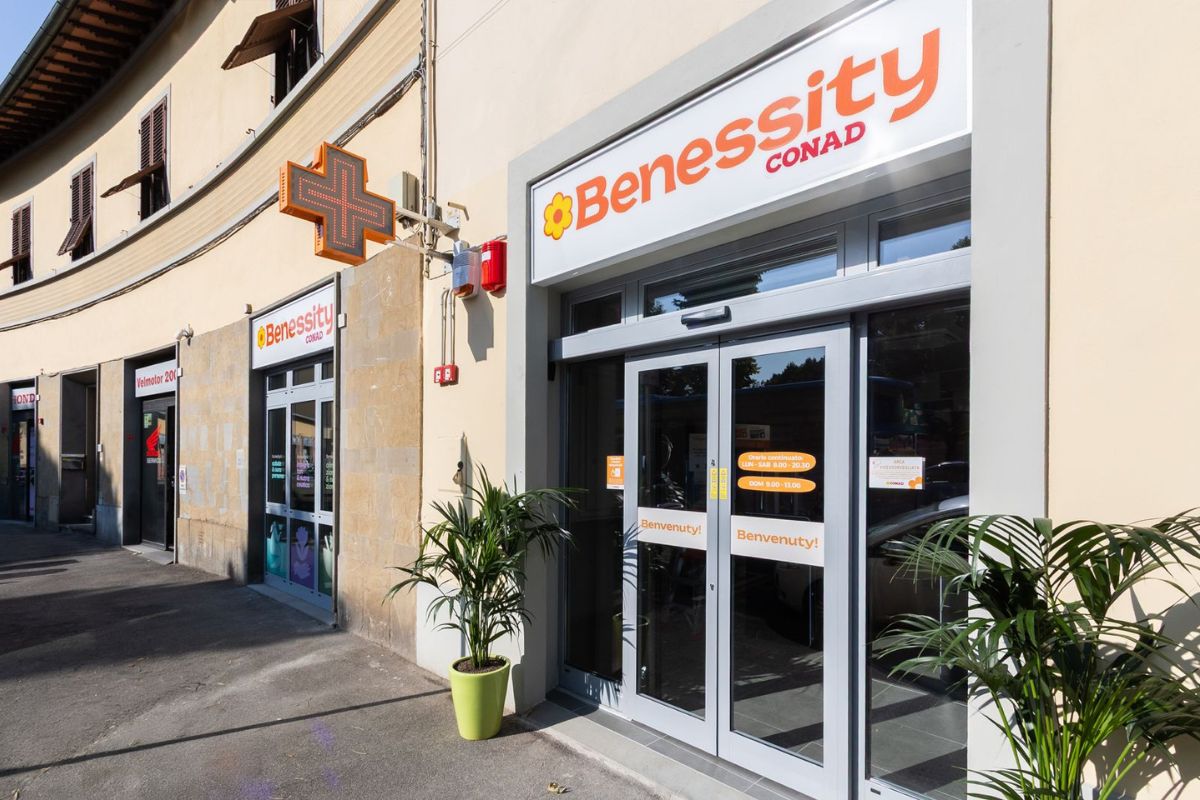 Ingresso della parafarmacia Benessity Conad con croce luminosa, vetrate e piante all'esterno del punto vendita.
