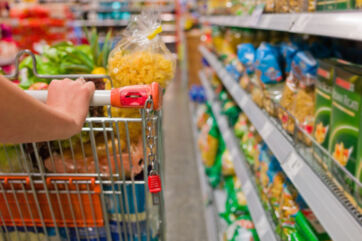 Mano spinge carrello della spesa nel supermercato tra scaffali di pasta e prodotti alimentari confezionati