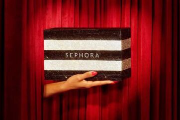 Mano che regge una scatola Sephora glitterata a righe su sfondo di tende rosse per il social commerce su TikTok.